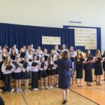 Grupa dzieci i dorosłych śpiewa na sali gimnastycznej. Ubrani w białe i czarne stroje, stoją przed niebieską kurtyną. Na ścianie widoczne są plakaty z napisami.