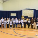 Grupa ludzi stoi w sali gimnastycznej na tle granatowych zasłon i plakatów. Przód ozdobiony jest dużymi, żółtymi kwiatami. Niektórzy trzymają dyplomy oraz torby.