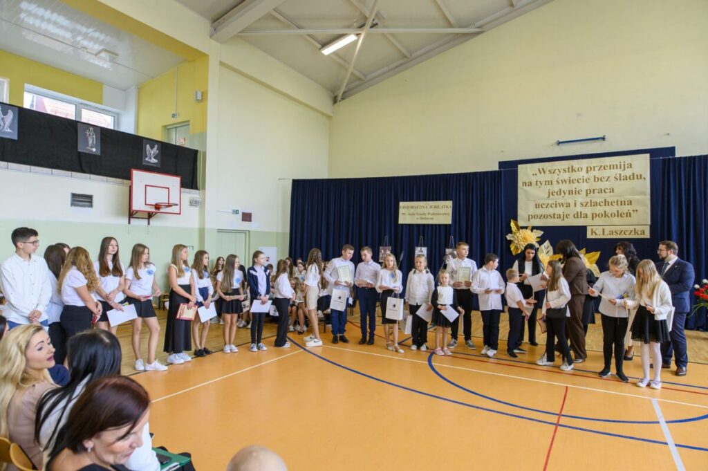 Grupa uczniów w strojach galowych stoi na drewnianej sali gimnastycznej. Tło ozdabiają niebieskie zasłony i cytat na plakacie. Uczniowie trzymają dyplomy i torby na nagrody.