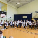 Grupa uczniów w strojach galowych stoi na drewnianej sali gimnastycznej. Tło ozdabiają niebieskie zasłony i cytat na plakacie. Uczniowie trzymają dyplomy i torby na nagrody.