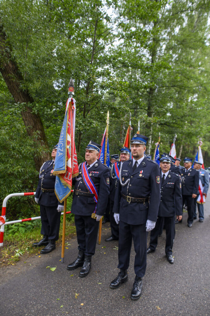 Grupa strażaków w mundurach stoi z flagami na tle zielonego lasu. Mężczyźni mają poważne wyrazy twarzy. Droga jest asfaltowa, z białymi i czerwonymi barierkami po lewej.