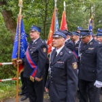 Grupa strażaków w mundurach z barwnymi sztandarami stoi na tle zielonego lasu. Dominuje kolor granatowy i czerwony. Tło jest pełne drzew i liści.