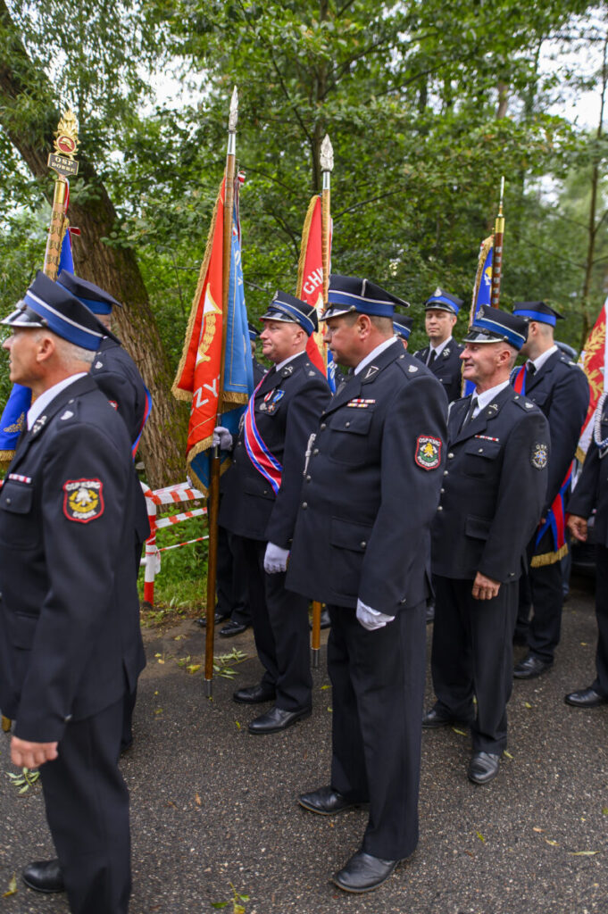 Grupa strażaków w mundurach stoi z kolorowymi sztandarami. Tło to zielone drzewa. Ujęcie z uroczystości plenerowej.
