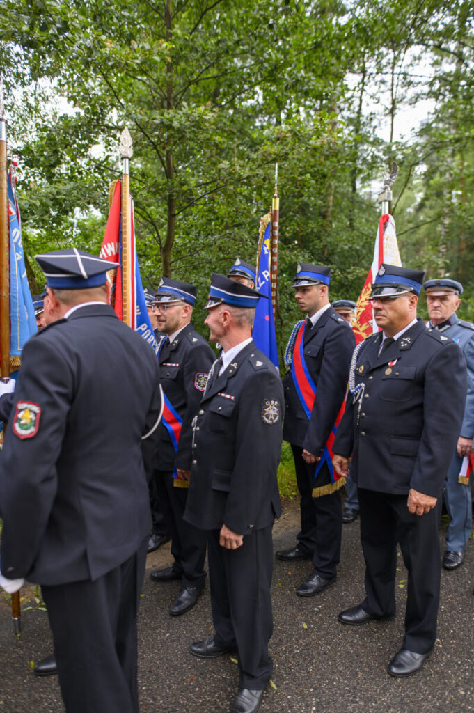 Grupa mężczyzn w mundurach strażackich z flagami stoi na tle zieleni. Mają na sobie ciemne uniformy z niebieskimi elementami. Tło stanowią drzewa z gęstymi liśćmi.