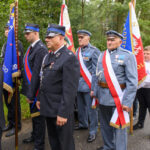 Grupa mężczyzn w mundurach stoi na ulicy z flagami w tle. Mundury są w kolorach czarnym i niebieskim. W tle jest zielony las.