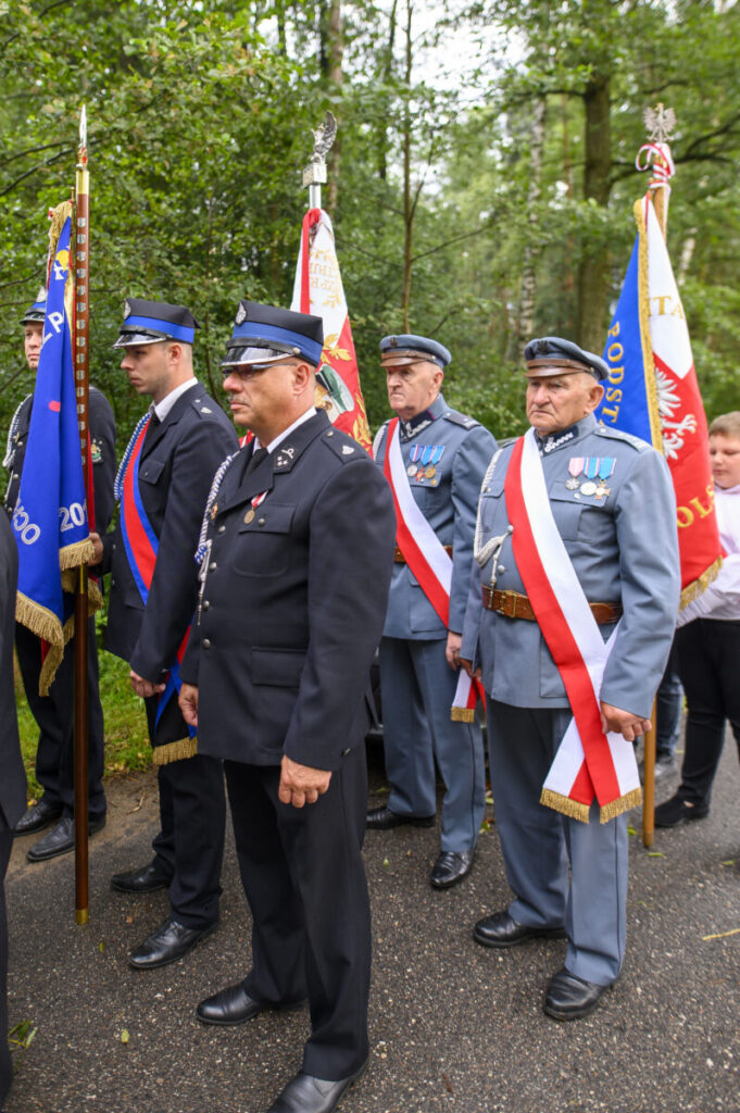 Grupa mężczyzn w mundurach stoi na ulicy z flagami w tle. Mundury są w kolorach czarnym i niebieskim. W tle jest zielony las.