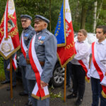 Na zdjęciu widać dwóch starszych mężczyzn w mundurach, z medalami, trzymających flagi. Tło stanowi zalesiony teren. Obok nich stoją młodsi chłopcy w białych koszulach z szarfami.