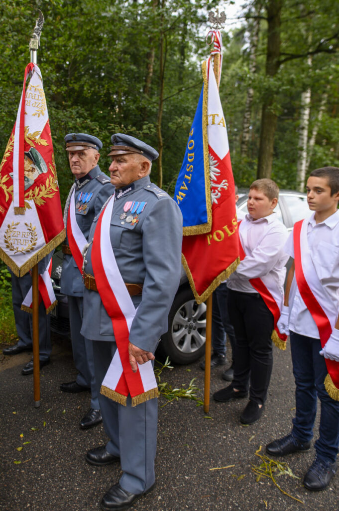 Na zdjęciu widać dwóch starszych mężczyzn w mundurach, z medalami, trzymających flagi. Tło stanowi zalesiony teren. Obok nich stoją młodsi chłopcy w białych koszulach z szarfami.