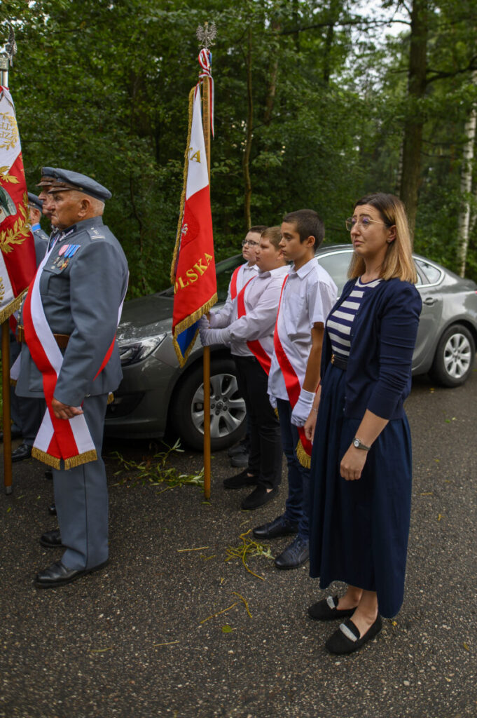 Grupa osób w odświętnych strojach stoi w szeregu z flagą Polski. Tło to zielony las. Przeważają kolory czerwony, biały i niebieski.
