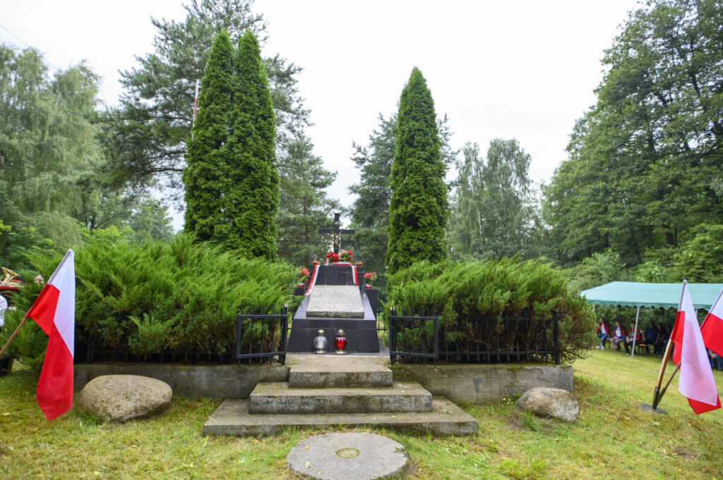 Cmentarz wojskowy z flagami Polski, otoczony zielenią. Pomnik w centrum z krzyżem i zniczami. Tło leśne.