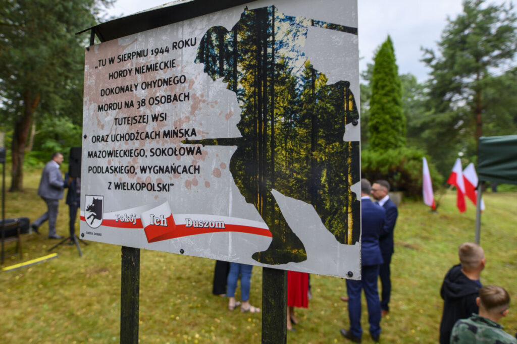 Tablica upamiętniająca wydarzenia z sierpnia 1944, z tekstem i grafiką lasu. W tle drzewa i ludzie w formalnych ubraniach. Obok powiewają flagi Polski.