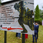 Tablica upamiętniająca wydarzenia z sierpnia 1944, z tekstem i grafiką lasu. W tle drzewa i ludzie w formalnych ubraniach. Obok powiewają flagi Polski.