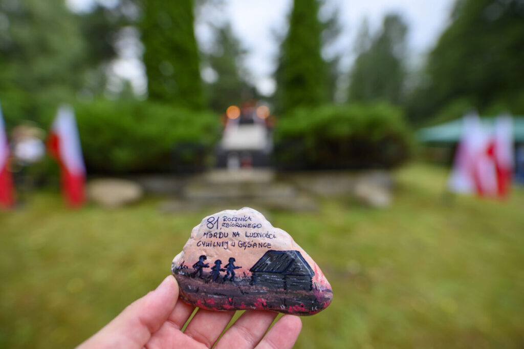 Ktoś trzyma pomalowany kamień z napisem i rysunkami. W tle rozmyte polskie flagi i zielone krzewy. Kolory: zielony, biały, czerwony.