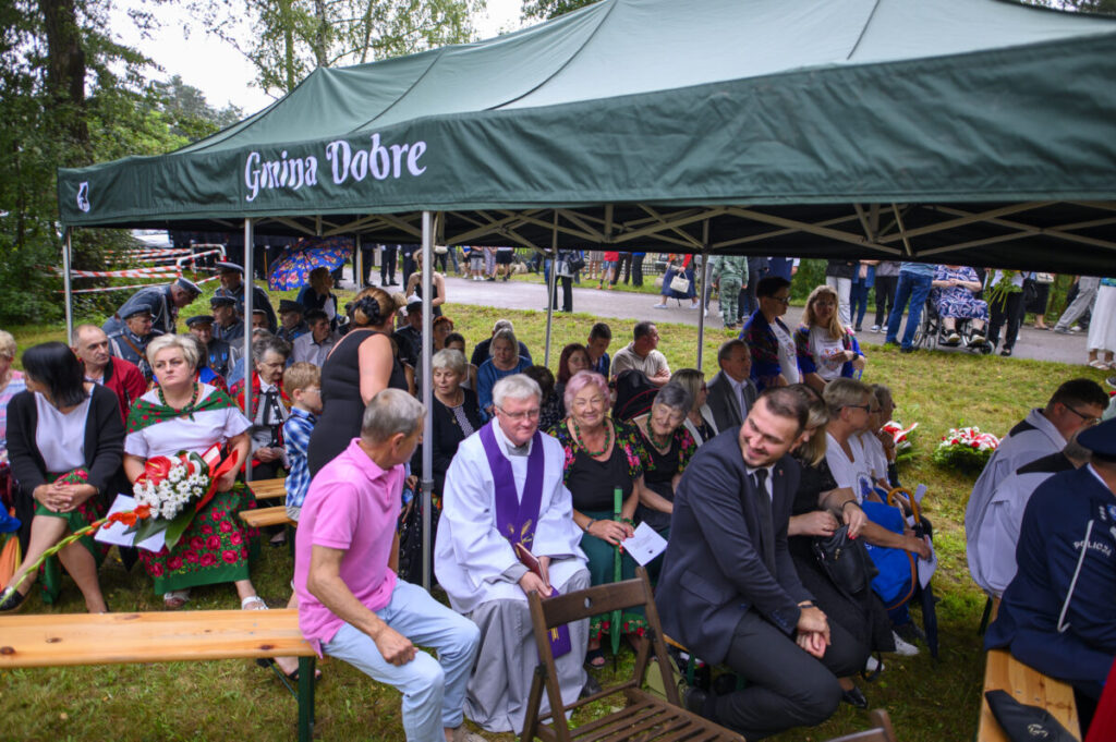 Grupa ludzi pod zielonym namiotem z napisem Gmina Dobre, siedzą na ławkach, tło stanowi zieleń drzew i trawa.