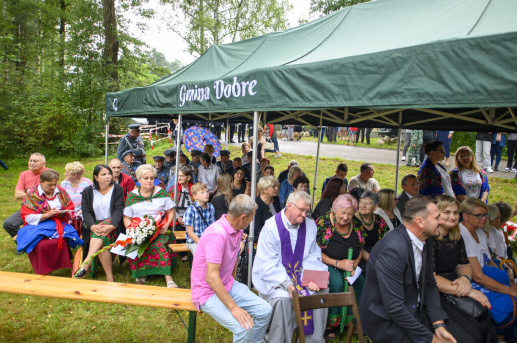 Grupa ludzi siedzi pod zielonym namiotem z napisem Gmina Dobre na tle drzew. Mają na sobie kolorowe ubrania. Pogoda pochmurna.
