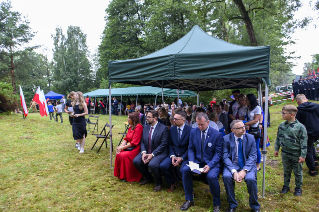 Na zdjęciu widać ludzi siedzących pod zielonym namiotem na tle lasu. W tle stoją trzy polskie flagi i gromadzą się inni uczestnicy wydarzenia. Trawa jest soczyście zielona, a uczestnicy noszą eleganckie stroje.