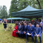 Na zdjęciu widać ludzi siedzących pod zielonym namiotem na tle lasu. W tle stoją trzy polskie flagi i gromadzą się inni uczestnicy wydarzenia. Trawa jest soczyście zielona, a uczestnicy noszą eleganckie stroje.