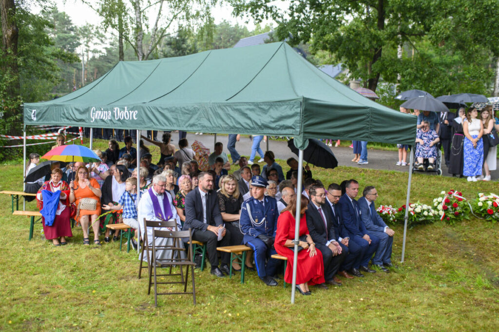Pod namiotem z napisem Gmina Dobre siedzą ludzie na ławkach. Tło to zielone drzewa, a niektorzy trzymają parasole. Kwiaty ułożone na trawie.
