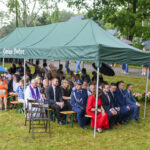 Pod namiotem z napisem Gmina Dobre siedzą ludzie na ławkach. Tło to zielone drzewa, a niektorzy trzymają parasole. Kwiaty ułożone na trawie.