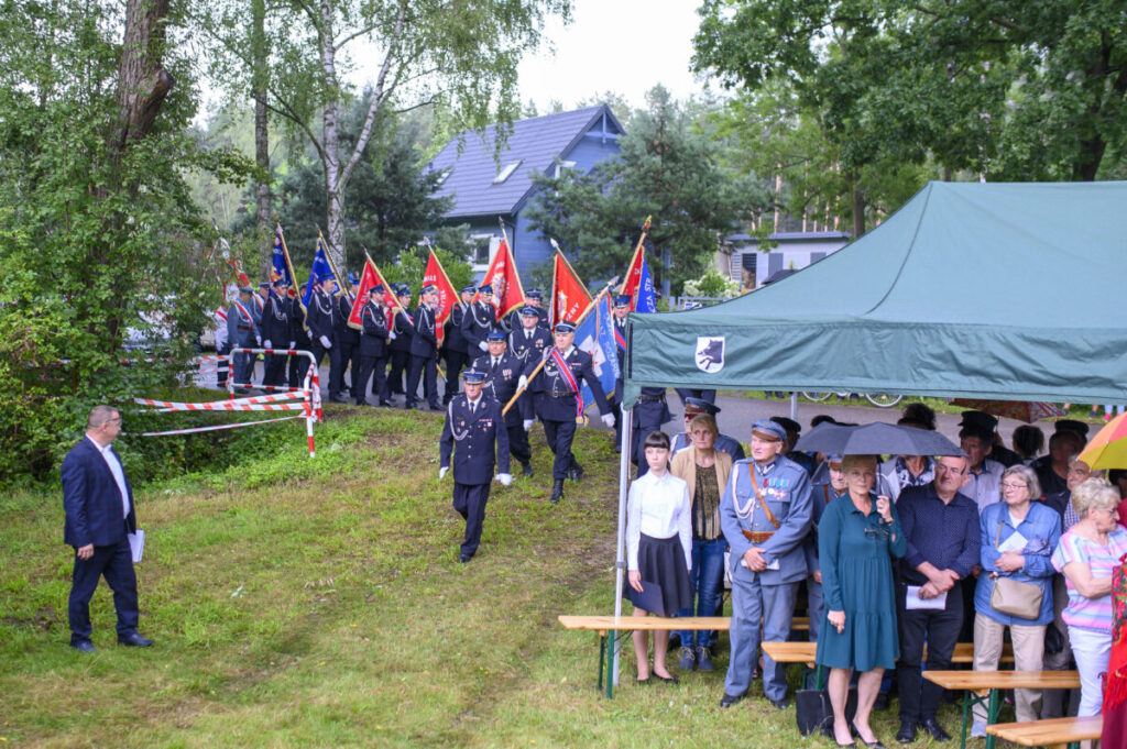 Na zdjęciu grupa ludzi w mundurach z flagami maszeruje przez zielony teren. Po prawej stronie stoi zielony namiot z osobami pod nim. W tle drzewa i dom.