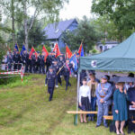 Na zdjęciu grupa ludzi w mundurach z flagami maszeruje przez zielony teren. Po prawej stronie stoi zielony namiot z osobami pod nim. W tle drzewa i dom.