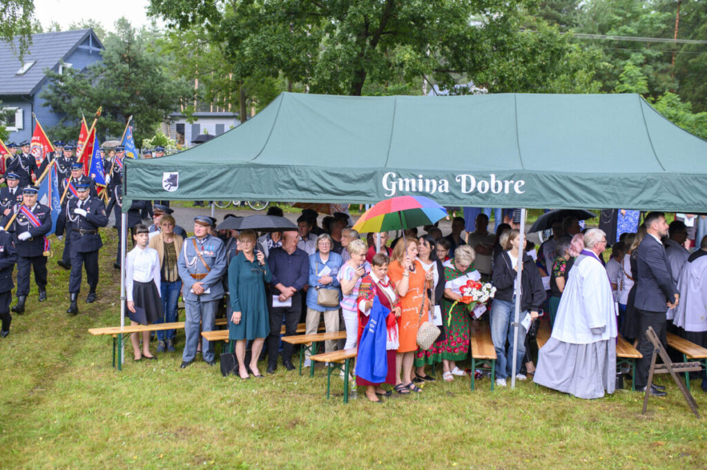 Grupa ludzi pod zielonym namiotem z napisem „Gmina Dobre”. Deszcz, kolorowe parasole. W tle drzewa i budynki.