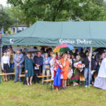 Grupa ludzi pod zielonym namiotem z napisem „Gmina Dobre”. Deszcz, kolorowe parasole. W tle drzewa i budynki.