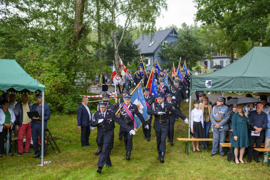 Grupa strażaków w mundurach maszeruje z flagami. Tło stanowią zielone drzewa i niebieski budynek. Po bokach zielone namioty z widzami.