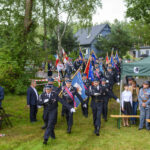 Grupa strażaków w mundurach maszeruje z flagami. Tło stanowią zielone drzewa i niebieski budynek. Po bokach zielone namioty z widzami.
