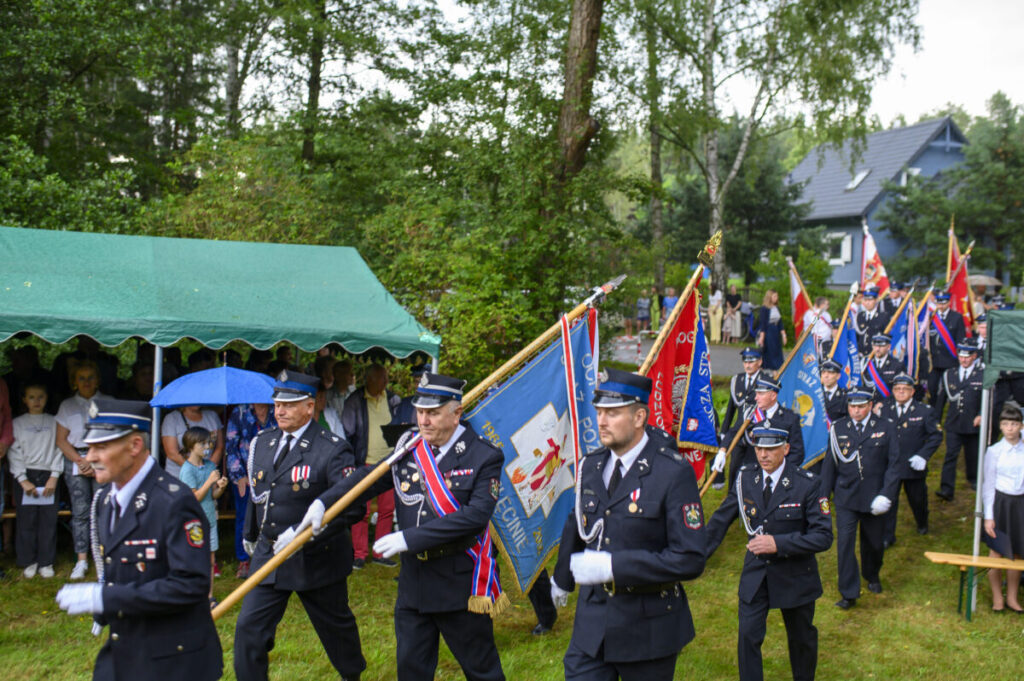 Grupa strażaków w paradzie niesie kolorowe flagi. Mundury są granatowe, a tło zielone od drzew. Widać namiot i widzów.