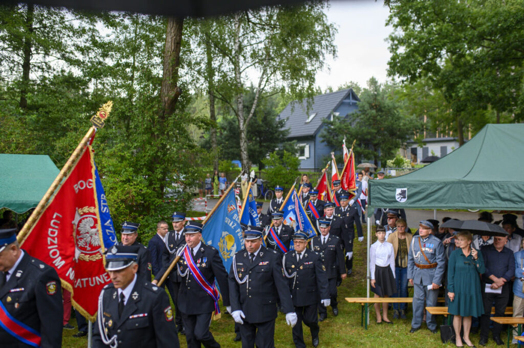 Grupa strażaków w mundurach niesie kolorowe sztandary, stojąc obok zielonego namiotu. W tle drzewa i dom. Uroczystość na świeżym powietrzu.