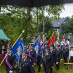 Parada strażaków w ciemnych mundurach maszerujących z niebieskimi i czerwonymi sztandarami. Tło: zielone drzewa, namioty, ludzie. Uroczystość pasuje do otoczenia w plenerze.