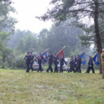 Grupa ludzi maszeruje w deszczu z flagami na tle zielonego lasu. Ubrani są w mundury, a obok stoi osoba salutująca. Kolory są stonowane, dominują zielenie i szarości.