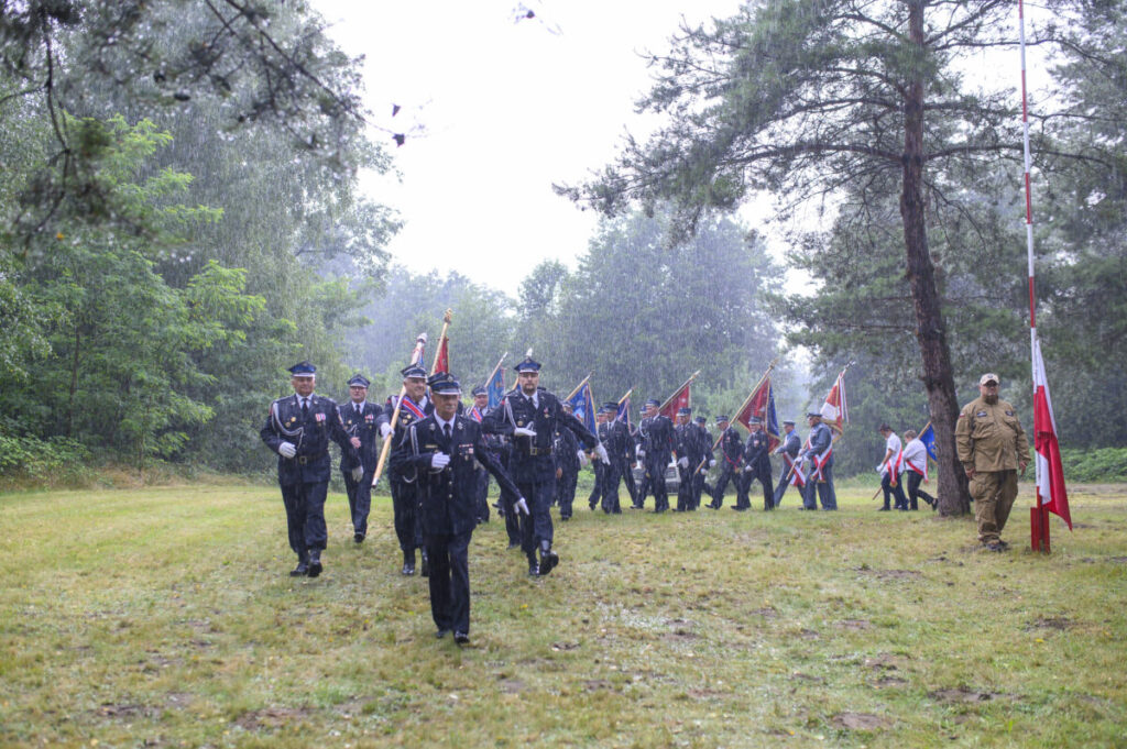 Grupa ludzi w mundurach maszeruje w deszczu z flagami w rękach. Tło to zielone drzewa i trawa. Obok stoi osoba przy masztu z biało-czerwoną flagą.