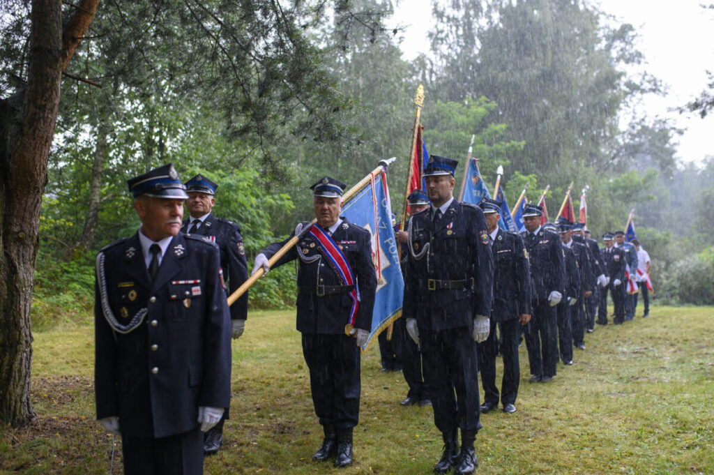 Grupa strażaków w mundurach maszeruje w deszczu. Każdy z nich trzyma kolorowy sztandar. W tle zieleń drzew.