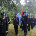 Grupa strażaków w mundurach maszeruje w deszczu. Każdy z nich trzyma kolorowy sztandar. W tle zieleń drzew.