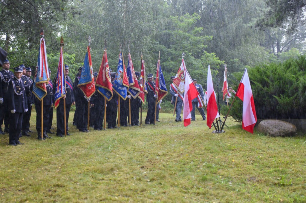 Grupa osób w mundurach trzyma kolorowe sztandary, stojąc na deszczu. W tle zielone drzewa. Z boku biało-czerwone flagi.