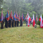 Grupa osób w mundurach trzyma kolorowe sztandary, stojąc na deszczu. W tle zielone drzewa. Z boku biało-czerwone flagi.