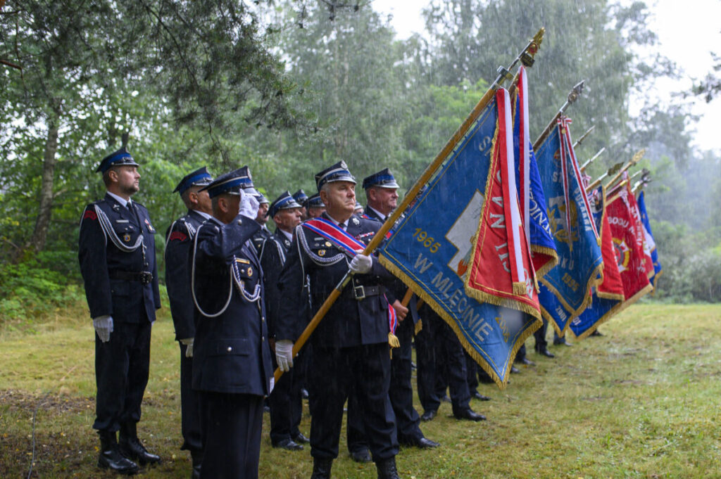 Grupa strażaków w mundurach stoi w deszczu na trawie z kolorowymi sztandarami. Tło to zielone drzewa. Uroczysta atmosfera.
