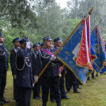 Grupa strażaków w mundurach stoi w deszczu na trawie z kolorowymi sztandarami. Tło to zielone drzewa. Uroczysta atmosfera.