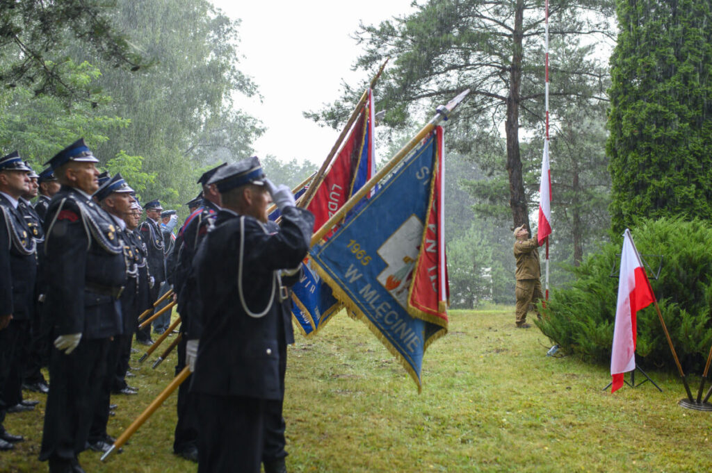 Grupa mundurowych w deszczu salutuje przy flagach. Tło z zielonymi drzewami. Centralnie unoszona biało-czerwona flaga.