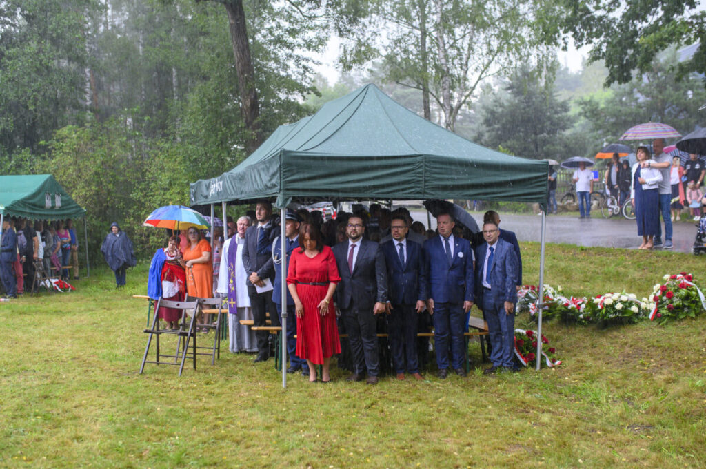 Ludzie stoją pod zielonym namiotem w deszczu, niektórzy trzymają parasole. Tło to drzewa i trawnik. Przód ozdobiony kwiatami.