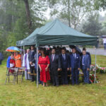 Ludzie stoją pod zielonym namiotem w deszczu, niektórzy trzymają parasole. Tło to drzewa i trawnik. Przód ozdobiony kwiatami.