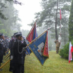 Grupa strażaków w czarnych mundurach trzyma kolorowe flagi podczas deszczu. W tle wysokie drzewa. Po prawej widoczny żołnierz przy maszcie z flagą.