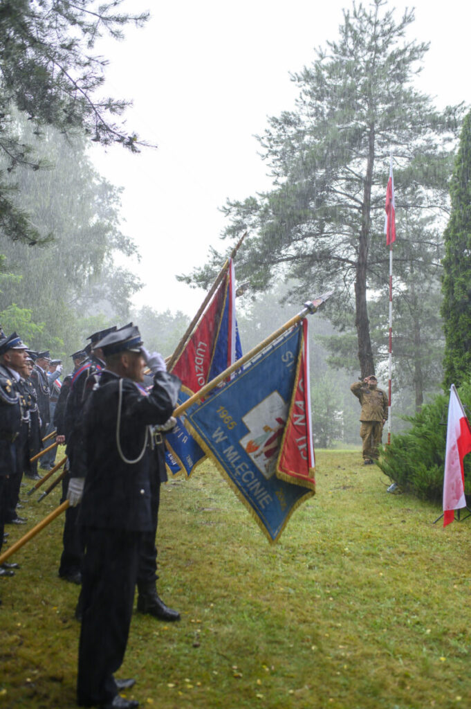 Grupa strażaków w czarnych mundurach trzyma kolorowe flagi podczas deszczu. W tle wysokie drzewa. Po prawej widoczny żołnierz przy maszcie z flagą.