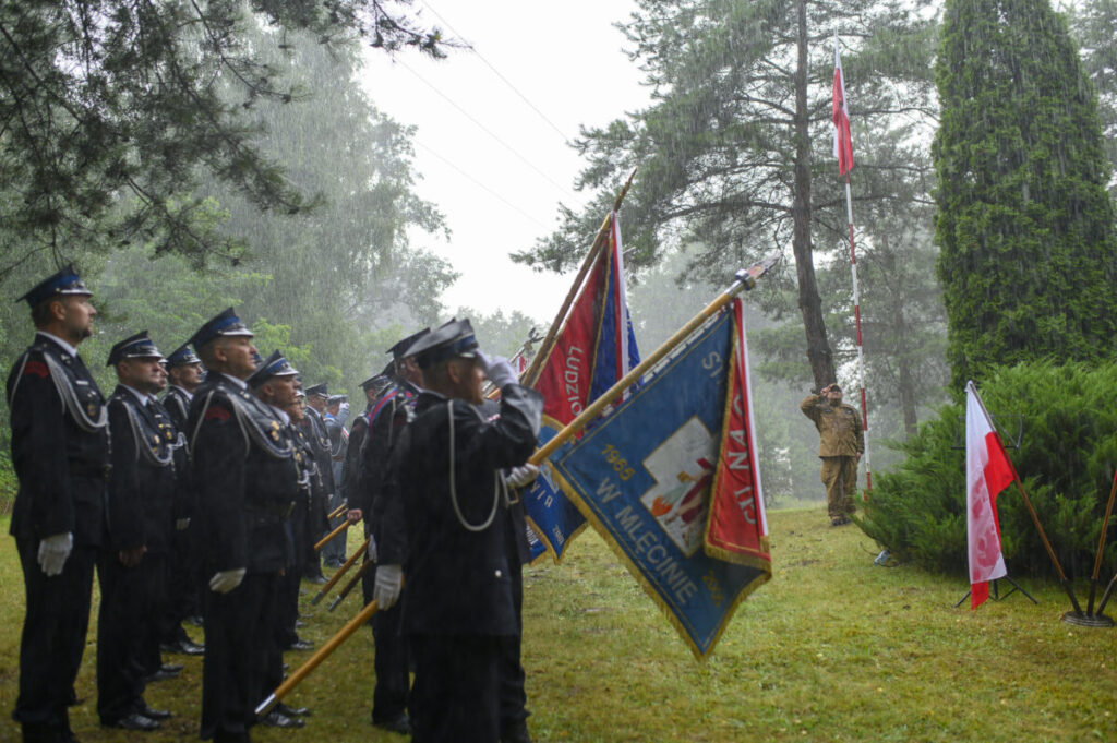 Grupa ludzi w mundurach trzymająca sztandary stoi na deszczu. Tło obrazują drzewa i polska flaga. Uroczystość ma charakter oficjalny.