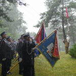Grupa ludzi w mundurach trzymająca sztandary stoi na deszczu. Tło obrazują drzewa i polska flaga. Uroczystość ma charakter oficjalny.