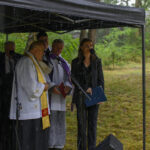Pod namiotem podczas deszczu stoi grupa osób w strojach liturgicznych i formalnych, mówiących do mikrofonów. Tło leśne.