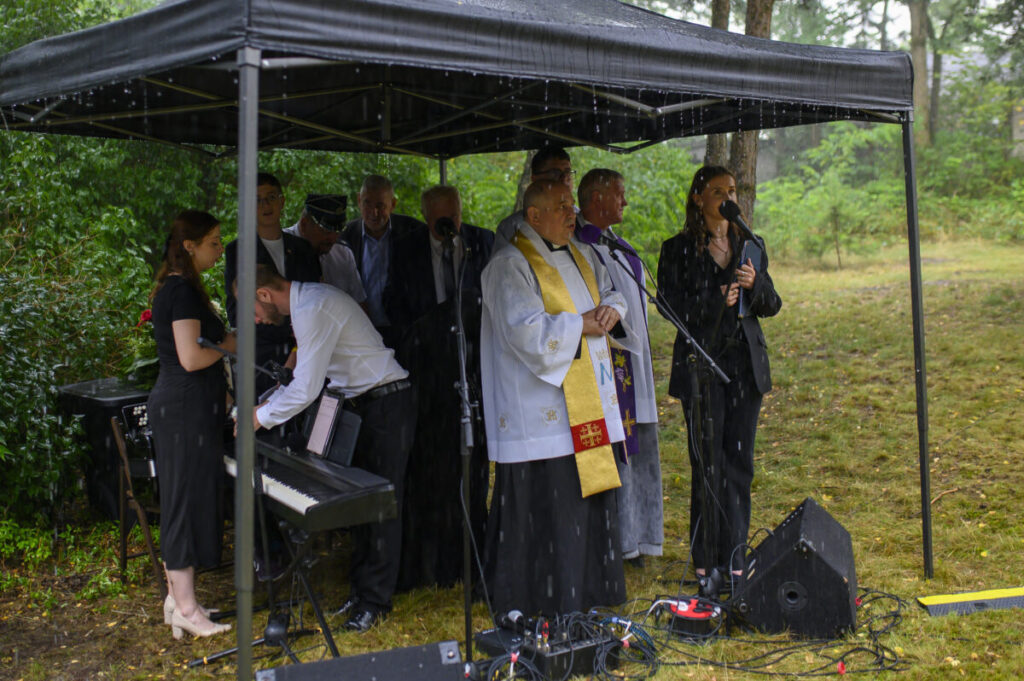 Grupa ludzi pod czarnym namiotem, podczas deszczu. Na pierwszym planie duchowny w stroju liturgicznym. Tło stanowi zielona roślinność.