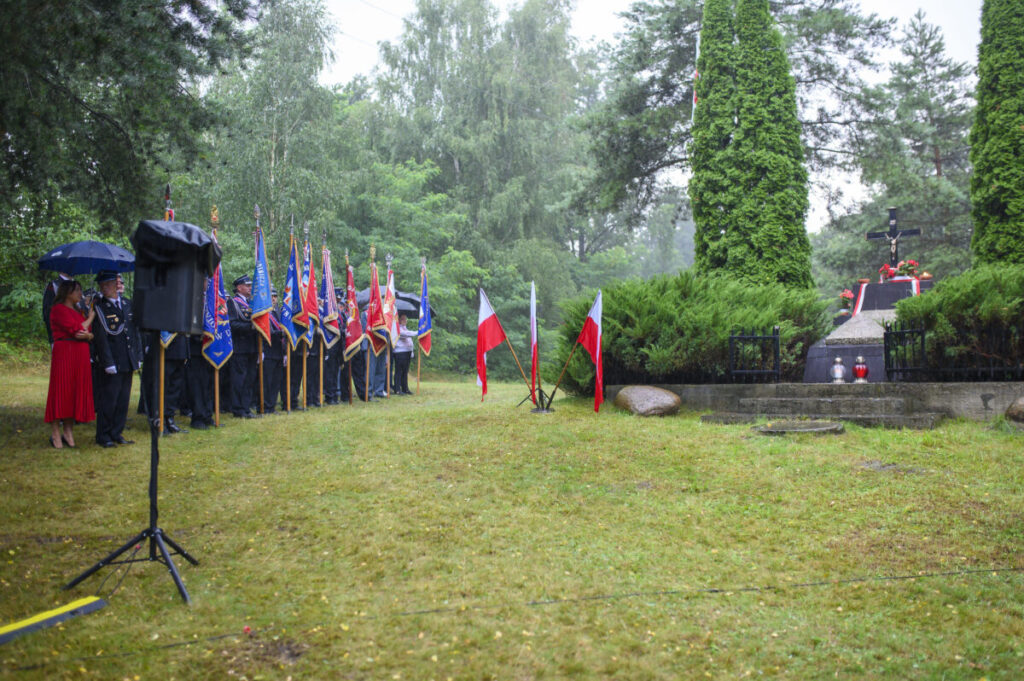 Grupa ludzi z flagami stoi na trawniku, w tle drzewa. Obok flagi Polski, widoczny jest pomnik z krzyżem. Uroczystość odbywa się w deszczu.
